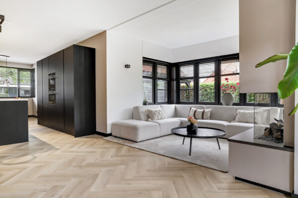 Albendo, vloeren specialist - Moderne woonkamer met Plak PVC Visgraat Premium Oak Beige - Dryback LV1260V vloeren, groot grijs bankstel, ronde zwarte salontafel, neutrale inrichting, hoge zwarte kasten en grote ramen met jaloezieën die uitkijken op groen.
