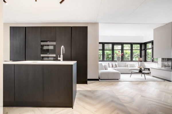 Albendo, vloeren specialist - Een moderne, open keuken met donkere kasten en een ingebouwde oven loopt over in een lichte woonkamer met lichte banken, grote ramen en een salontafel. Plak PVC Visgraat Premium Eik Beige - Dryback LV1260V vloeren verenigen de ruimte.