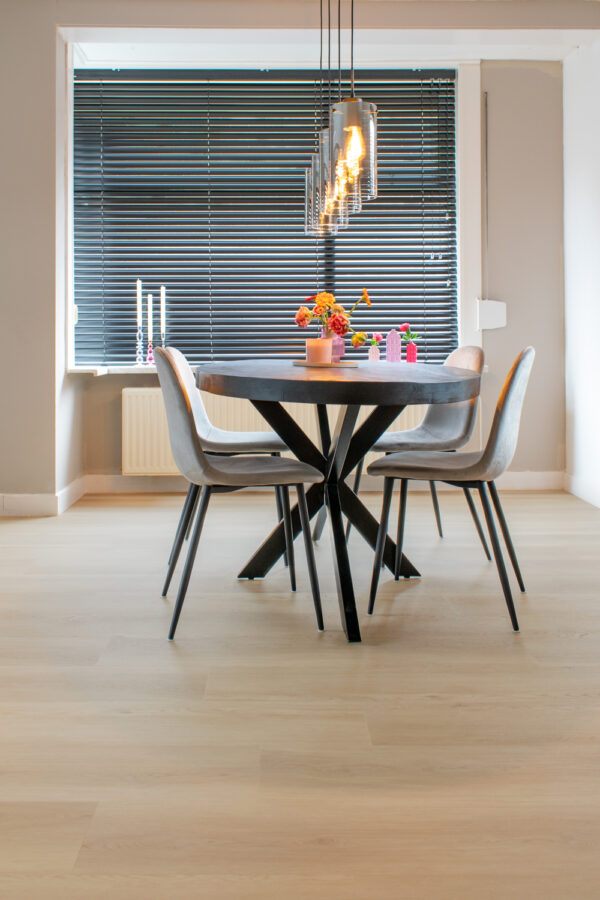 Albendo, vloeren specialist - Een moderne eetkamer heeft een ronde tafel, vier grijze stoelen, hangende verlichting en zwarte jaloezieën. Bloemen en kaarsen sieren de tafel, die op een Plak PVC Beige Extra Matte Toplaag - Dryback LV2300P vloer ligt.