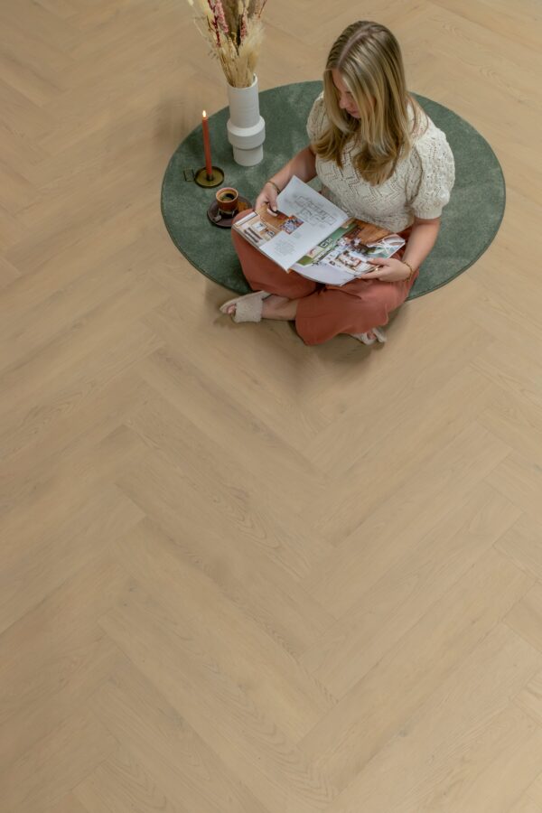 Albendo, vloeren specialist - Een vrouw zit in kleermakerszit op een groen cirkelvormig tapijt op een Plak PVC Visgraat Natural Light - Dryback LV1902V vloerbedekking en leest een tijdschrift. Naast haar staan een brandende kaars, een schaaltje en gedroogde bloemen.