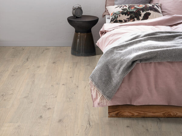 Albendo, vloeren specialist - Een gezellige slaapkamerhoek heeft een bed met roze beddengoed, een deken met grijs patroon en een gebloemd kussen. Een rond zwart bijzettafeltje met een klokje staat op een elegante Laminaat Kasteel Eiken vloer.