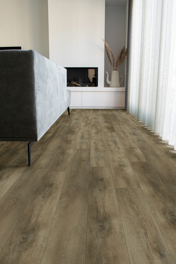Albendo, vloeren specialist - Een moderne woonkamer is voorzien van Plak PVC Valley Oak Smoked - Drayback LV6000P vloeren, een grijze bank, een witte kast met gedroogd pampagras in een vaas, een open haard en een raam met transparante gordijnen die natuurlijk licht binnenlaten.