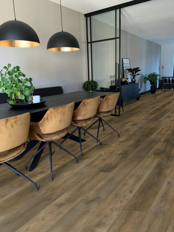 Albendo, vloeren specialist - Een moderne eetkamer met een lange zwarte tafel, bruine fluwelen stoelen, twee zwarte hanglampen, groene planten en Plak PVC Valley Oak Pepper - Dryback LV6001P vloeren. Grote glazen panelen en een neutrale inrichting versterken de eigentijdse stijl.
