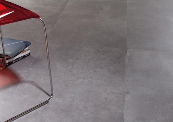 Albendo, vloeren specialist - Een close-up van de grijze tegelvloer Nexus Antraciet Mat 90x90cm met een rode stoelpoot en een stapel tijdschriften gedeeltelijk zichtbaar in de linkerbovenhoek.
