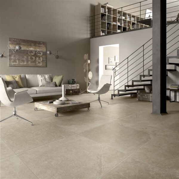Albendo, vloeren specialist - Een moderne woonkamer met neutrale tinten, Vloertegel / Wandtegel Mainz Beige 90x90cm vloertegels, een grijze bank, twee witte stoelen, een lage salontafel en een metalen trap die leidt naar een open zolderruimte met planken en boeken.