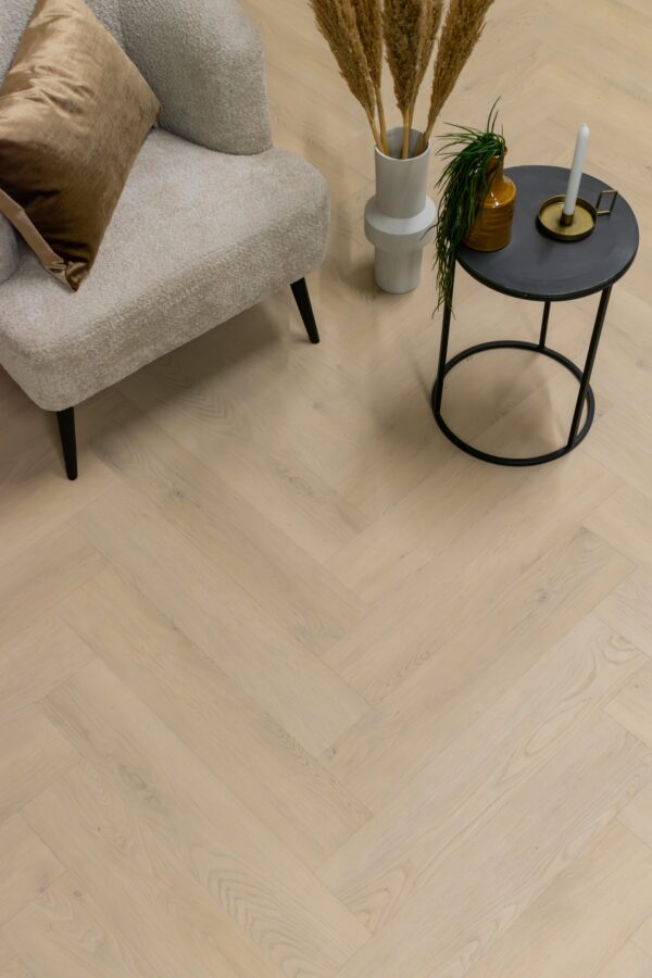 Albendo, vloeren specialist - Een gezellige beige fauteuil met een bruin kussen staat naast een klein zwart tafeltje met decor, allemaal geplaatst op Click PVC Visgraat 2901 (harde klik) Beige vloeren in een licht visgraatpatroon.