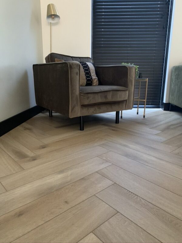Albendo, vloeren specialist - Een moderne woonkamerhoek met een fauteuil van bruin fluweel, een geometrisch kussen van zwart en goud, een plantje op een bijzettafeltje en een Click Visgraat 5201 (harde klik) vloer van natuurlijk eiken licht. Zwarte jaloezieën bedekken het raam.