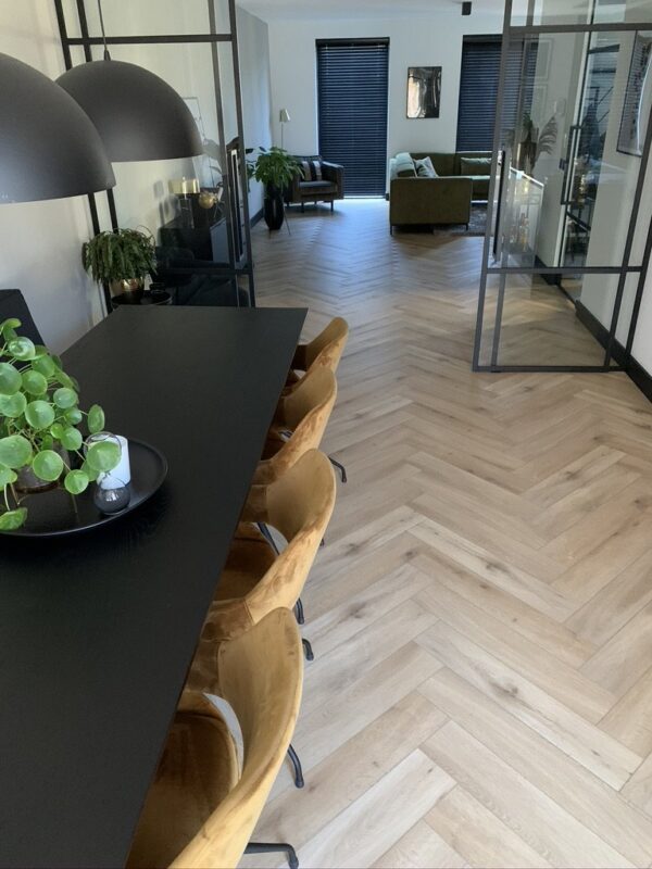 Albendo, vloeren specialist - Moderne eetkamer met een zwarte tafel, gele fluwelen stoelen, kamerplanten en hanglampen; natuurlijk eiken licht klik Visgraat 5201 vloer leidt naar een woonkamer met groene banken en zwarte vensterrolgordijnen op de achtergrond.