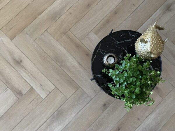 Albendo, vloeren specialist - Op een ronde zwarte tafel staan een groene potplant, een kaars en een gouden vissculptuur, allemaal op de Click Visgraat 5201 (rigide click) Natural Oak Light vloer.