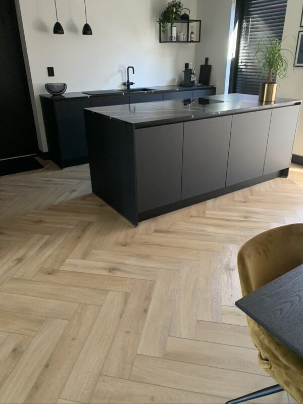 Albendo, vloeren specialist - Moderne keuken met donkere kasten, eiland, matzwarte armaturen en minimalistische inrichting. De vloer is Click Visgraat 5201 (harde klik) Eik natuur licht in een visgraatmotief. Erboven hangen hanglampen; in de hoek staan een gele stoel en tafel.