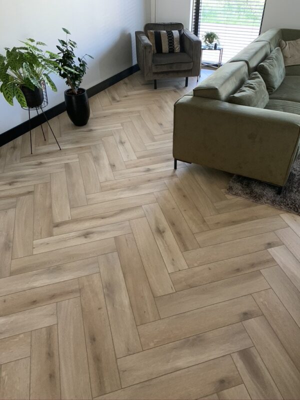 Albendo, vloeren specialist - Moderne woonkamer met Click Visgraat 5201 (harde klik) Natural Oak Light vloeren, een groene sofa, grijze fauteuil met kussen, twee potplanten en een raam met horizontale jaloezieën die natuurlijk licht binnenlaten.
