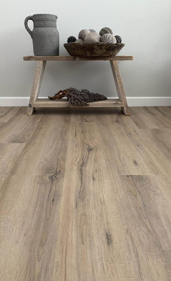 Albendo, vloeren specialist - Een klein houten tafeltje met een kannetje en een schaaltje staat tegen een bleke muur op een vloer met Click PVC 5600 (rigid click) Smoked Oak Light, die scherp in beeld is terwijl de tafel en decoratieve voorwerpen een beetje wazig op de achtergrond lijken.