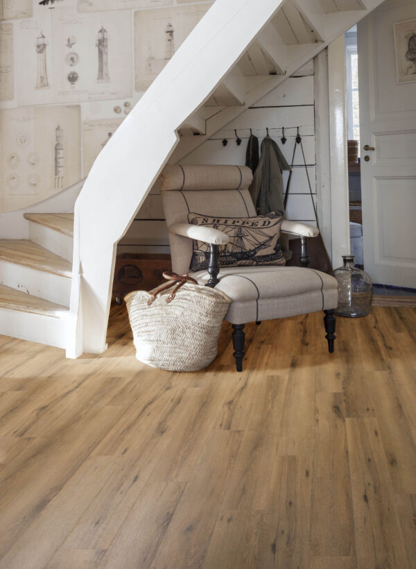 Albendo, vloeren specialist - Een gezellige leeshoek onder een trap is voorzien van een beige fauteuil met kussens met patroon, een geweven mand, muurhaken met jassen, nautische muurkunst en een vloer met houtlook in Click PVC 5601 (harde klik) Gerookt Eiken Natuur.