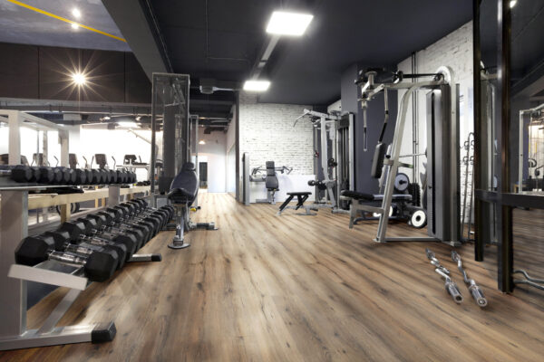 Albendo, vloeren specialist - Een moderne fitnesszaal met Click PVC 5601 (harde klik) vloeren in gerookt eiken natuur, met vrije gewichten, banken, krachttoestellen, spiegelende muren en heldere bovenverlichting voor een schone en ruime uitstraling.