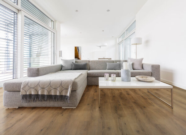 Albendo, vloeren specialist - Een moderne woonkamer heeft een grote grijze bank, een kleed met patroon, een witte salontafel met decor en grote ramen met jaloezieën. De vloer is bedekt met Click PVC 5601 (harde klik) gerookt eiken naturel.