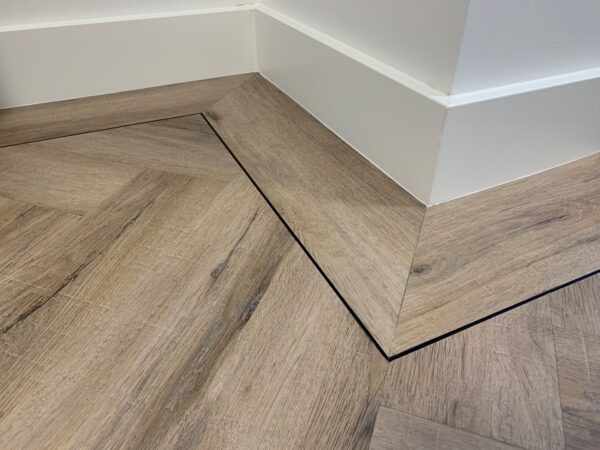 Albendo, vloeren specialist - Close-up van een kamerhoek met Click Visgraat 5700 (harde klik) gerookt eiken licht vinyl vloeren met witte plinten. De schuine planken passen precies in de hoek en benadrukken een geometrisch patroon.