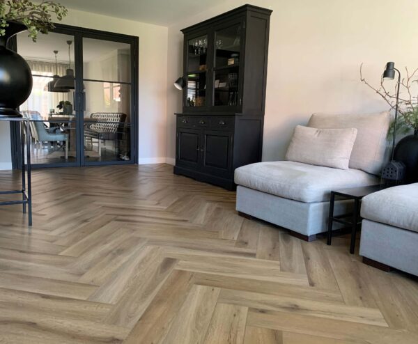 Albendo, vloeren specialist - Een moderne woonkamer met Click Visgraat 5202 (harde klik) visgraatvloer in natuurlijk eiken, een grote grijze stoel met kussens, een zwarte kast en glazen deuren die leiden naar een eethoek met een tafel en stoelen.