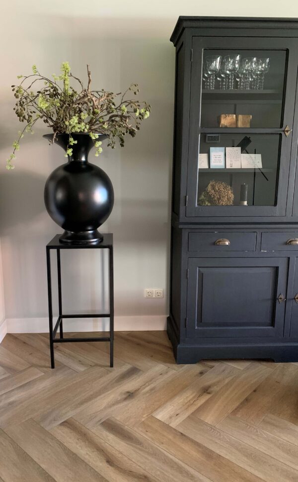 Albendo, vloeren specialist - Een zwarte vaas met groene takken op een hoge standaard staat naast een zwarte kast met glazen deuren waarop decor staat, allemaal op een Click Visgraat 5202 (stijve klik) naturel eiken vloer tegen een lichtgrijze muur.
