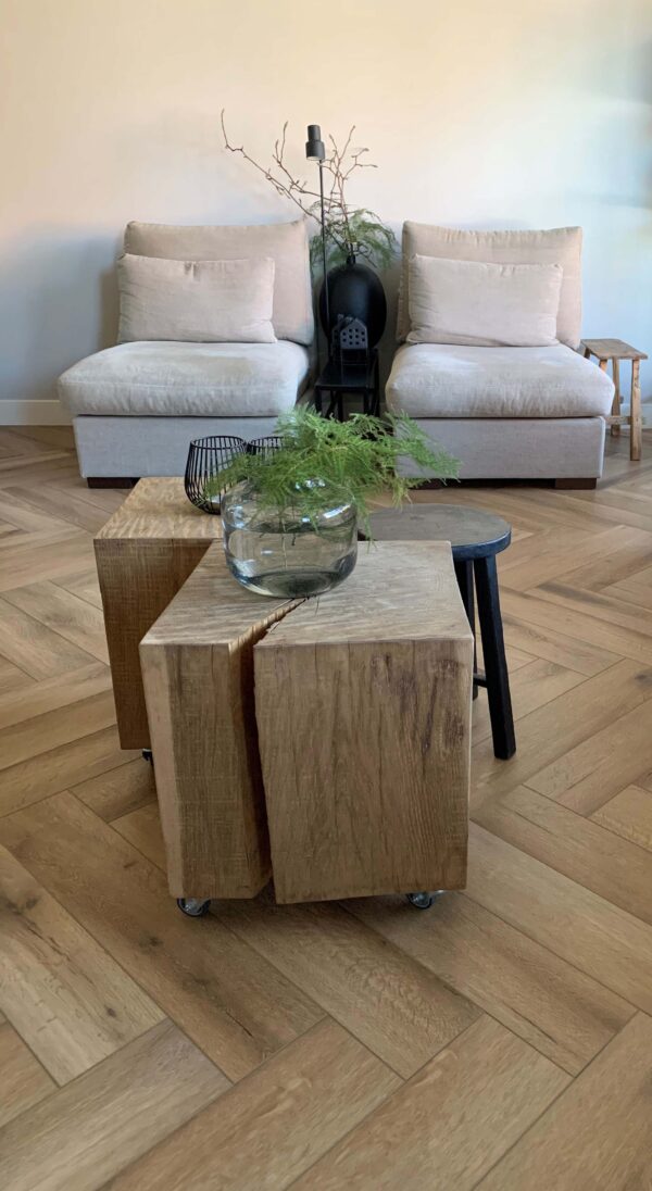 Albendo, vloeren specialist - Twee beige loungestoelen staan tegen een effen muur, met aan de voorkant een rustieke houten salontafel met een glazen schaal met groen en een mand van zwart draad. De kamer heeft een minimalistische inrichting en een Click Visgraat 5202 (harde klik) visgraatvloer van natuurlijk eiken.