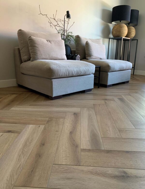Albendo, vloeren specialist - Twee beige loungestoelen met grote kussens staan tegen een lichte muur. Een lamp, vaas en decor staan op een tafeltje vlakbij. Op de vloer ligt Click Visgraat 5202 (harde klik) Natuurlijk Eiken in een visgraatpatroon.