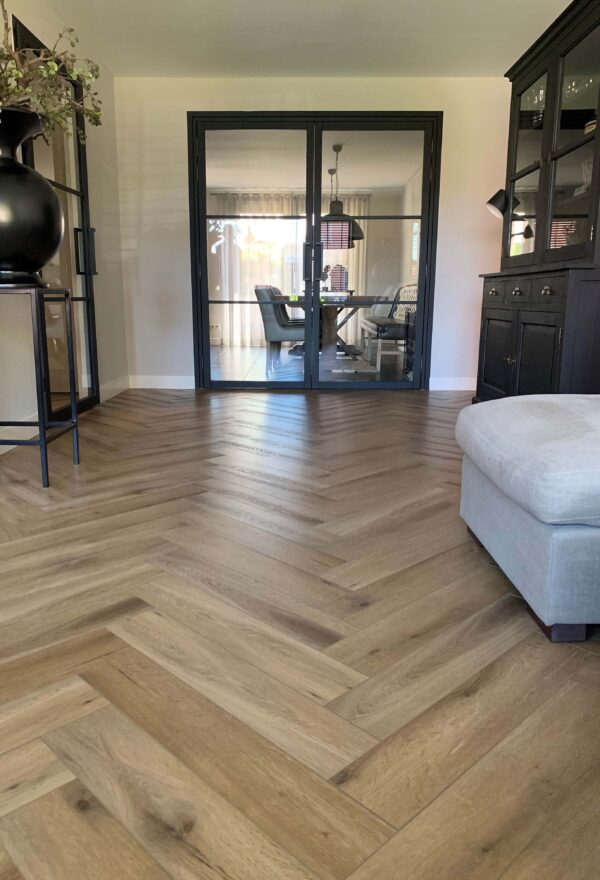 Albendo, vloeren specialist - Een moderne woonkamer is voorzien van Click Visgraat 5202 (harde klik) visgraatvloer in natuurlijk eiken, dubbele deuren in zwart omlijst glas en zwart meubilair. Een lichtbeige voetenbank staat op de voorgrond met een zithoek achter de deuren.