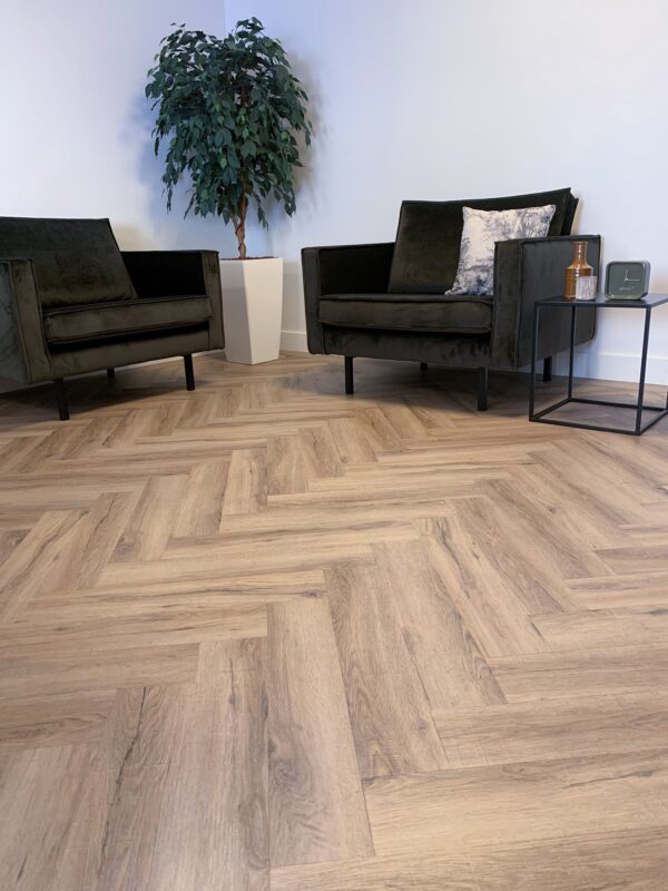 Albendo, vloeren specialist - Een moderne kamer met Click Visgraat 5700 (harde klik) Smoked Oak Light visgraatvloer, twee donkere fauteuils met kussens, een potplant in de hoek en een kleine bijzettafel met een fles, glas en decoratief object.