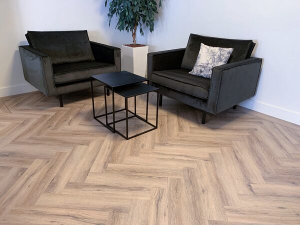 Albendo, vloeren specialist - Twee donkere fauteuils met gedessineerde kussens staan tegenover elkaar op zwarte nesttafels op een Click Visgraat 5700 (harde klik) visgraatvloer in gerookt eiken licht. Een hoge groene potplant staat in de hoek tegen een witte muur.