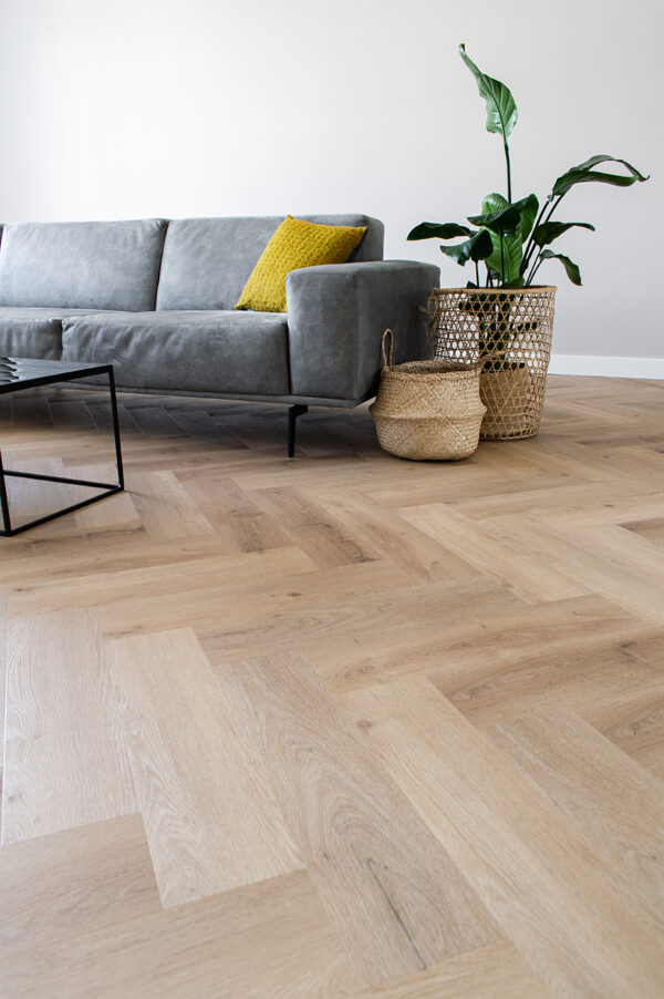 Albendo, vloeren specialist - Een moderne woonkamer met Click PVC Visgraat 5400 (Rigid Click) Prestige Oak Natural visgraatvloer, een grijze bank met een geel kussen, zwarte metalen salontafel, plant in pot, twee gevlochten manden en lichtgekleurde muren voor een lichte uitstraling.