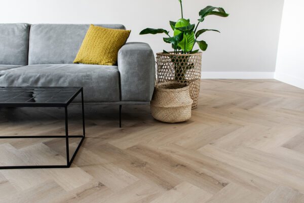 Albendo, vloeren specialist - Een moderne woonkamer met een grijze bank, geel kussen, zwarte metalen salontafel, twee gevlochten manden en een groene potplant op Click PVC Visgraat 5400 (Rigid Click) Prestige Oak Natural visgraatvloer.