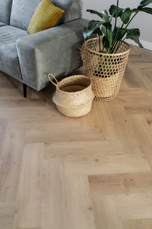 Albendo, vloeren specialist - Een grijze bank met een geel kussen staat naast een groene plant in pot en twee gevlochten manden op een Prestige Oak Natural Click PVC Visgraat 5400 (Rigid Click) vloer in een moderne woonruimte.