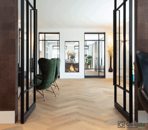 Albendo, vloeren specialist - Moderne leefruimte met Click PVC Visgraat 5400 (Rigid Click) Prestige Eik Natuur visgraatvloer, groen fluwelen fauteuils, glazen deuren met zwart frame en zicht op een open haard en bloemstuk op de achtergrond.