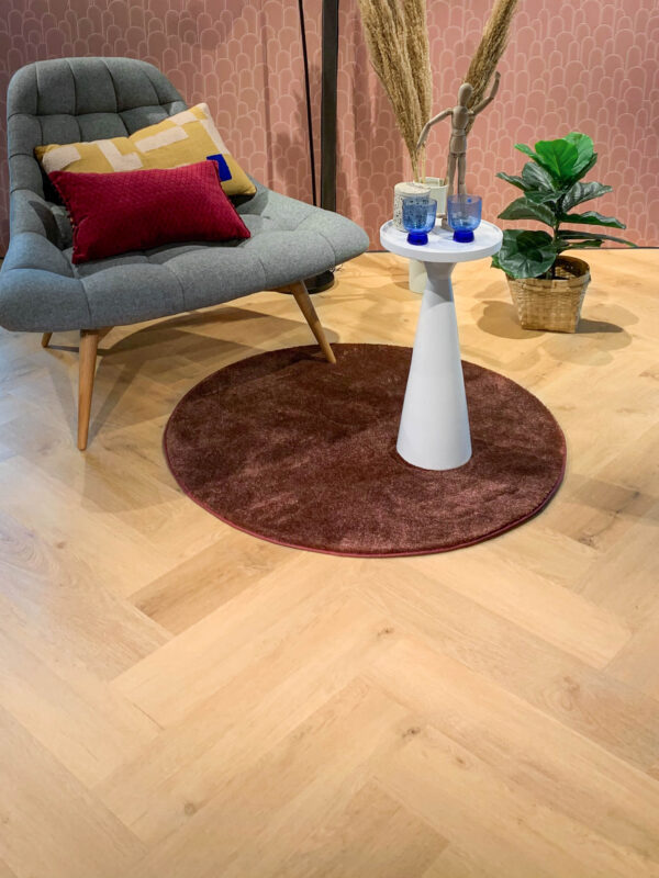 Albendo, vloeren specialist - Een moderne lounge is voorzien van een grijze accentstoel, kleurrijke kussens, een kleine witte ronde tafel met decor, een kastanjebruin cirkelvormig vloerkleed, een plant in pot, roze geometrisch behang en Prestige Oak Natural Click PVC Visgraat 5400 (Rigid Click) vloeren.