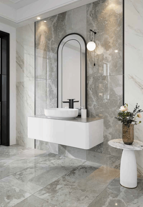 Albendo, vloeren specialist - Moderne badkamer met Kale Greige Matte 60x120cm vloer- en wandtegels, een zwevend wit badmeubel met wasbak, hoge gewelfde spiegel, zwarte kraan, wandlamp en een klein rond bijzettafeltje met een vaas bloemen.