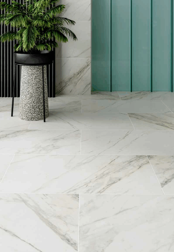 Albendo, vloeren specialist - Een modern interieur met Kale Apuano White 60x120cm vloertegels, een hoge groene plant in pot op een gespikkelde standaard, zwarte verticale wandpanelen en een blauw glazen wandpaneel op de achtergrond.