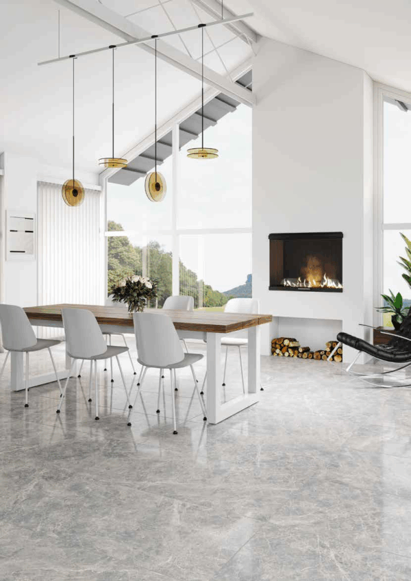 Albendo, vloeren specialist - Moderne eetkamer met Kale Corinthian Beige 60x120cm tegels, witte stoelen rond een houten tafel, gouden hanglampen, open haard aan de muur met netjes gestapeld haardhout, grote ramen en kamerplanten.