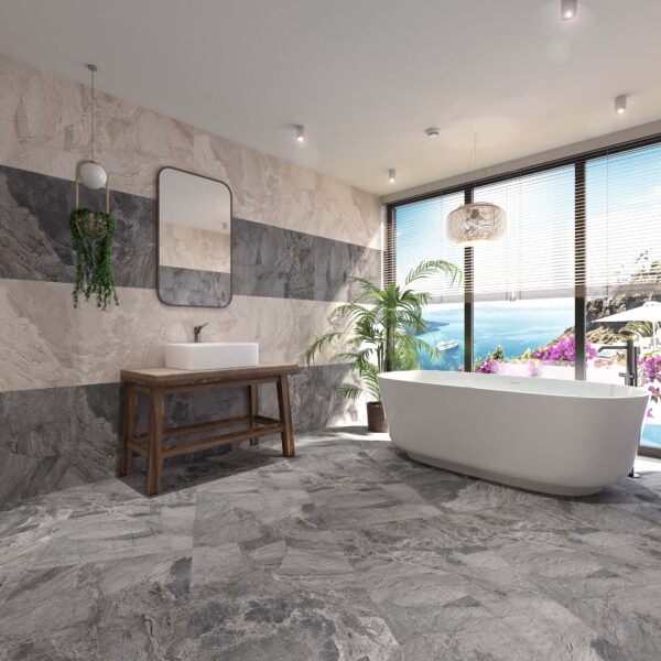 Albendo, vloeren specialist - Een moderne badkamer met Bodrum Silver 60x120cm grijze stenen tegels, een vrijstaand wit bad bij grote ramen met uitzicht op de oceaan, planten in potten, een houten badmeubel met wastafel en een hangende spiegel.